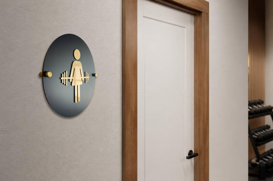 Black and gold women's restroom sign with a weightlifter icon on standoff mounts outside a gym locker room door