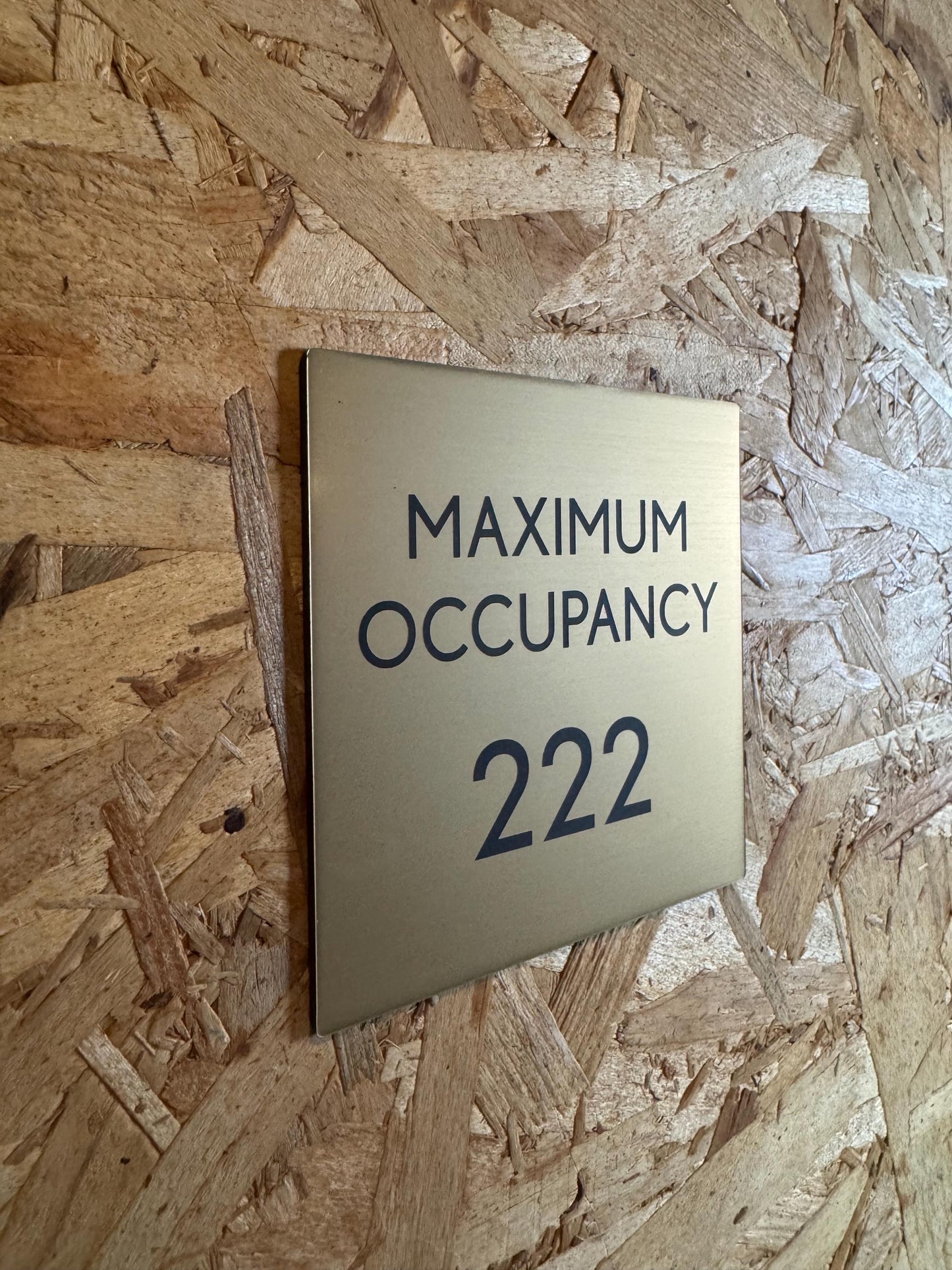 May include: A gold-colored metal sign with black text. The sign displays the words &#39;MAXIMUM OCCUPANCY&#39; above the number &#39;222&#39;. The sign is mounted on a light brown wood-like surface.