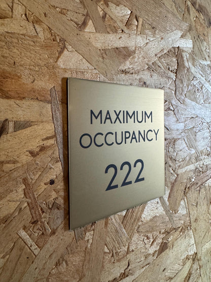 May include: A gold-colored metal sign with black text. The sign displays the words &#39;MAXIMUM OCCUPANCY&#39; above the number &#39;222&#39;. The sign is mounted on a light brown wood-like surface.