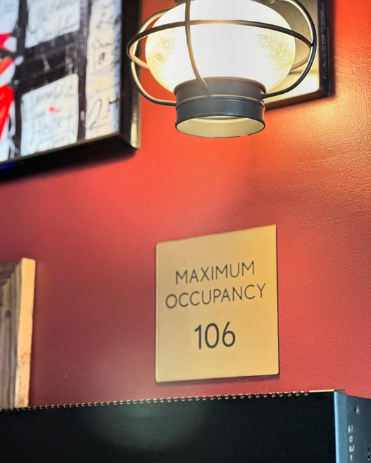 May include: Close-up view of a gold-colored metal sign on a red wall. The sign displays the text &#39;MAXIMUM OCCUPANCY&#39; above the number &#39;106&#39;.  A dark-colored metal cage light fixture with a frosted glass globe is partially visible in the upper left corner of the image.  The wall is a deep reddish-brown.  The sign is rectangular with rounded corners.